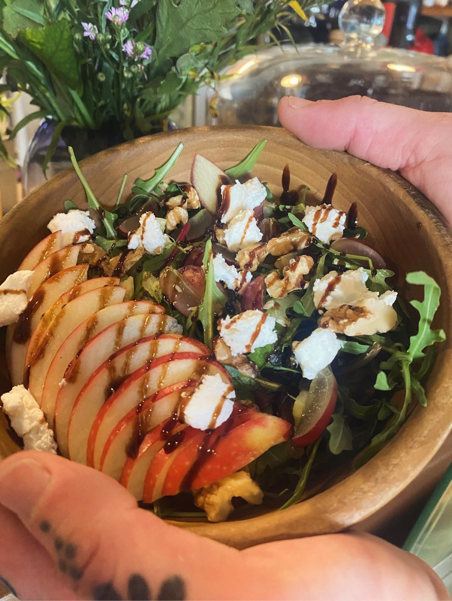 Two hands holding a wooden bowl of apple, goat's cheese, grape and walnut salad in the café