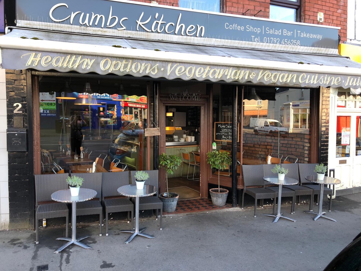 The Crumbs Kitchen shopfront on Gwydr Square: grey awning, outdoor tables and bay trees either side of the door